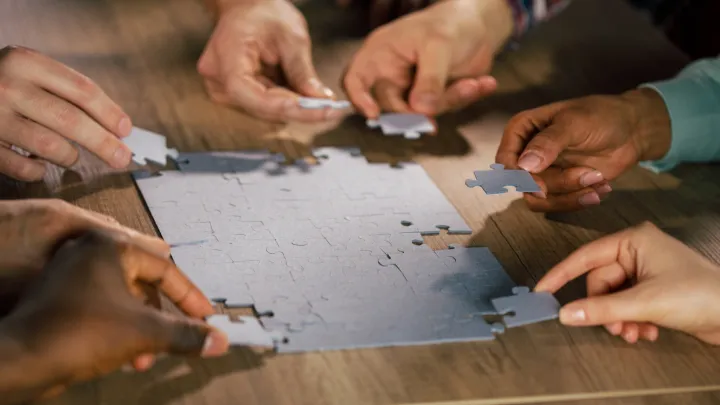 A close-up shot of multiple hands working together to assemble a jigsaw puzzle on a wooden table, symbolizing teamwork and collaboration.
